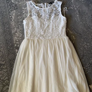 Vit spetsklänning med ärmlös design - Superfin vit klänning med spets upptill och ärmlös design. Kjolen är i flera lager tunt tyg som ger ett luftigt och romantiskt intryck. Perfekt för sommarens alla tillfällen och studentfirande. Dragkedja i ryggen för enkel påklädning.