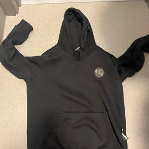 Svart hoodie från Philipp Plein - Svart hoodie från Philipp Plein med klassisk huva och magficka. Tröjan har ett stilrent märke på bröstet och är långärmad. Perfekt för dig som gillar en clean och snygg streetstil.