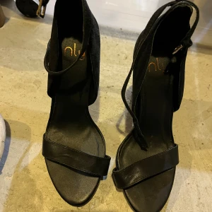 Svarta klackade sandaler från NLY - Svarta sandaler från NLY med hög klack och öppen tå. Skorna har en bred rem över tårna och en snygg ankelrem med spänne. Materialet är en mix av mocka och skinn, vilket ger en lyxig känsla. Perfekta för att lyfta din outfit.