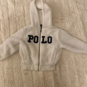 Vit fleecehoodie från Polo Ralph Lauren - Mysig vit hoodie från Polo Ralph Lauren i teddyfleece med broderad POLO-text framtill. Tröjan har dragkedja, huva och ribbade muddar vid ärmslut och midja. Perfekt för kyliga dagar och riktigt skön att ha på sig. Barn storlek