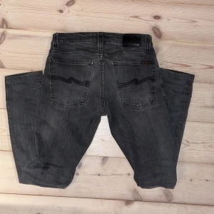 Grå jeans från Nudie Jeans Co W25 L32 - Säljer ett par grå jeans från Nudie Jeans Co i klassisk femficksmodell. Jeansen har svarta detaljer på bakfickorna och svart läderpatch i midjan. Modellen är slim fit och materialet är mjukt denim i bomull. Perfekta för dig som gillar en stilren look.