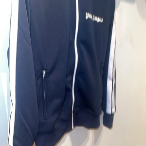 Palm Angels marinblå track jacket - Palm Angels marinblå track jacket med vita ränder längs ärmarna och vit dragkedja. Jackan har broderad logga på bröstet, ribbade muddar och två fickor framtill. Tillverkad i polyester för en sportig vibe och perfekt passform.