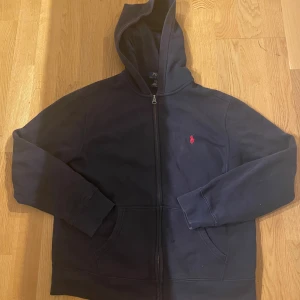 Blå hoodie från Polo Ralph Lauren - Marin Blå hoodie från Polo Ralph Lauren med dragkedja och huva. Klassisk röd broderad logga på bröstet och stora fickor framtill. Tillverkad i mjuk bomull, perfekt för chill dagar. Snygg och enkel att matcha med allt. Säljs pågrund av att den inte används längre. Sick 9/10 st160cm