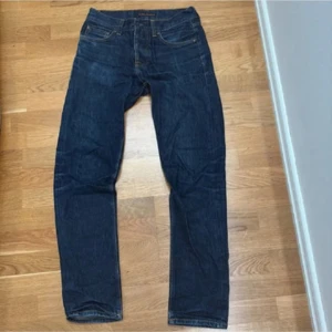 Mörkblå jeans från Nudie Jeans, W28 L32 - Säljer ett par mörkblå jeans från Nudie Jeans med klassisk femficksdesign och snygga kontrastsömmar. Jeansen har raka ben och normal passform, tillverkade i slitstarkt bomullsjeans. Bakfickorna har Nudie Jeans karaktäristiska broderi.