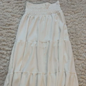 Vit långkjol med volanger - Säljer en vit långkjol med bred resår i midjan och flera volanglager som ger en flowig och chill vibe. Kjolen är i ett mjukt och lätt material, perfekt för varma dagar. Enkel att matcha med crop tops eller stickade tröjor. Använd max 3 ggr.