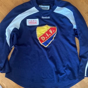 Blå långärmad DIF fotbollströja Reebok - Träningströja A-lag från 2005. Säljer en blå långärmad träningströja från Reebok med stort DIF-märke på bröstet och vita detaljer längs axlar och ärmar. Tröjan har broderad Reebok-logga och sponsorpatch på bröstet. Perfekt för dig som hejar på Djurgården.