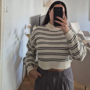 Randig stickad tröja - Säljer en stickad tröja i vitt med svarta horisontella ränder. Tröjan har hög krage, långa ärmar och ribbade muddar. Perfekt för dig som gillar en trendig stil.