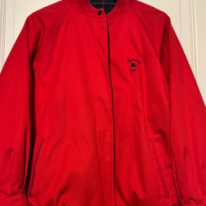 Vintage Burberrys Sport jacka röd/rutig – vändbar 2-i-1  - Exklusiv vändbar (2-i-1) vintagejacka från Burberrys Sport, tillverkad i England under sent 1980-tal eller tidigt 1990-tal. Jackan kan bäras åt båda hållen – ena sidan i klassiskt rött, och den andra i Burberrys rutmönstrade foder. Märkning: “Burberrys.London – REG 12 B87A” (autentisk vintageetikett). Burberrys London-etikett (med det äldre namnet “Burberrys”, som användes innan företaget tog bort ”s:et” i slutet av 1990-talet). PRIS KAN DISKUTERAS ❕❗️❕❗️