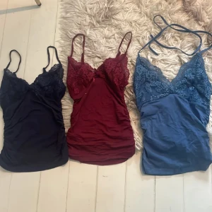 3 spetslinnetoppar i marinblå, vinröd och blå - Säljer tre snygga linnetoppar med spetsdetaljer i olika färger: marinblå, vinröd och blå. Alla har smala axelband och v-ringad spets upptill som ger en romantisk vibe. Perfekt att styla med jeans eller kjol för en trendig look.