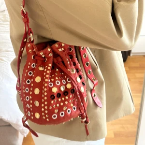 Marc by Marc Jacobs Tribal Pixie Bucket Bag - Sällsynt och karaktärsfull bucketväska från Marc by Marc Jacobs. Modellen Tribal Pixie Bucket Bag i korallrött äkta läder med dekorativa öljetter och studs i guld, silver, svart och vitt.  Tillverkad i läder med justerbar axelrem på cirka 115 cm. Väskan stängs med dragsko och har en mjuk, följsam struktur som ger en exklusiv känsla. Perfekt för vardag, resa eller som färgstark accent till en mer neutral outfit. Får plats med den nödvändigaste!   Väskan är i mycket fint skick 🤍