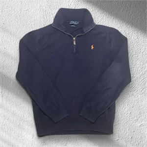 Marinblå half zip tröja Polo Ralph Lauren - Snygg marinblå tröja från Polo Ralph Lauren med klassisk orange logga på bröstet. Modellen har en half zip-dragkedja och hög krage. Tillverkad i mjuk pima bomull och har långa ärmar med ribbade muddar. Perfekt för en chill och stilren look.