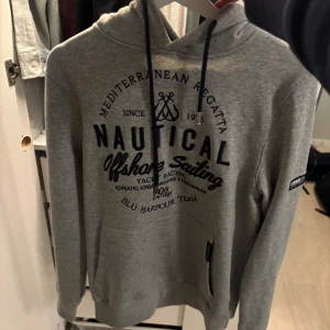 Grå hoodie från Blu Harbour - Snygg grå hoodie från Blu Harbour med marinblå tryck och snörning i huvan. Tröjan har känguruficka framtill och ribbade muddar vid ärmslut och nederkant. Perfekt för chill dagar och har en sportig vibe med nautiskt tema.