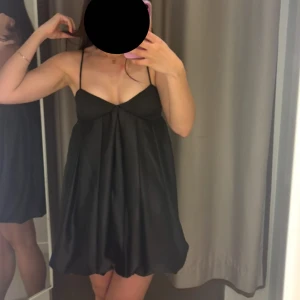 Svart kort klänning i satin med tunna axelband - HELT NY MED PRISLAPP.Supersöt svart kort klänning med tunna axelband och v-ringad urringning. Klänningen har en luftig, lätt utställd passform och är tillverkad i ett glansigt material som ger en elegant känsla. Perfekt för dig som gillar en stilren och chic look. Passar xs-m har resår på baksidan (se bild). Toppen har en pushup effekt på något vis