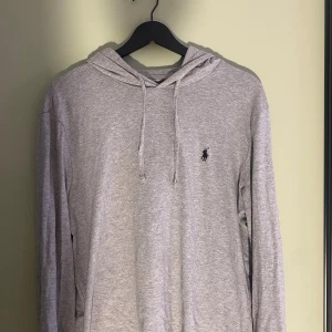 Grå hoodie från Polo Ralph Lauren - Snygg grå hoodie från Polo Ralph Lauren i mjuk bomull. Klassisk design med huva och snörning, samt broderad svart logga på bröstet. Perfekt för chill dagar och enkel att matcha med jeans.