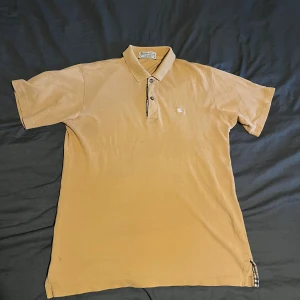 Beige pikétröja från Burberry Storlek S - Snygg beige pikétröja från Burberry med klassisk krage och knappslå. Tröjan har korta ärmar och diskret Burberry-logga på bröstet. Detaljer med rutigt tyg vid knappslå och nederkant ger en extra touch. Perfekt för dig som gillar stilrena och tidlösa plagg. Hör av er vid frågor!