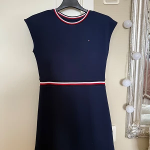 Marinblå klänning Tommy Hilfiger - Snygg marinblå klänning från Tommy Hilfiger med sportiga detaljer. Klänningen har ribbad halsringning med röd, vit och marinblå rand samt matchande rand vid midjan. Ärmlös modell med rak passform och diskret logga på bröstet. Perfekt för en stilren look.