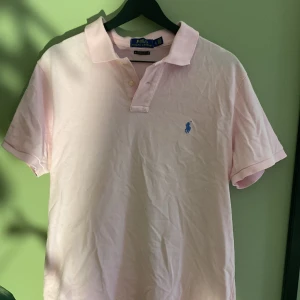 Rosa pikétröja från Polo Ralph Lauren - Snygg ljusrosa pikétröja från Polo Ralph Lauren med blå broderad logga på bröstet. Modellen är custom slim fit och har klassisk krage, korta ärmar och knappar framtill. Litet märke som jag visar i bilden
