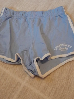 Blå shorts Los Angeles Beach H&M - Säljer ett par ljusblå shorts från H&M med vit kant och broderad text 'Los Angeles Beach' på benet. De har elastisk midja och är gjorda i 100% bomull, perfekta för chill dagar eller stranden. !!endast swish +frakt!!