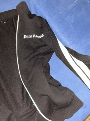 Söker byten eller säljer  - Säljer en svart hoodie från Palm Angels i storlek L. Tröjan har vit dragkedja, vita stripes längs ärmarna och Palm Angels-logga på bröstet. Klassisk streetwear-look med ribbade muddar och mjukt material. Perfekt för dig som vill ha en clean och stilren vibe.