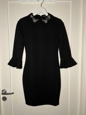 Svart kort klänning med pärlkrage - Supersnygg svart kort klänning från Amisu med trekvartsärm och volang vid ärmslut. Kragen är dekorerad med pärlor och stenar i blommönster som ger en extra touch. Figurnära passform och dragkedja bak. Perfekt för dig som vill sticka ut lite extra!