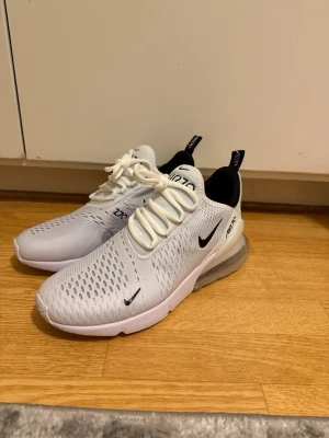 Nike Air Max 270 vita sneakers - Nike Air Max 270 sneakers i vitt med svarta detaljer och synlig luftbubbla i hälen. Ovandelen är i ventilerande syntetmaterial med snörning och svart Nike-logga på sidan. Snygg och sportig design som sticker ut med sin chunky sula och moderna look.