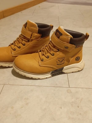 Dockers bruna boots med snörning - Snygga bruna boots från Dockers med snörning och kardborreband upptill. Skorna har en beige ovandel i mocka, mörkbrunt vadderat skaft och vit sula. Insidan är fodrad för extra värme och komfort. I färgen Tabaco. Perfekta för höst och vinter! Supersnygga! 