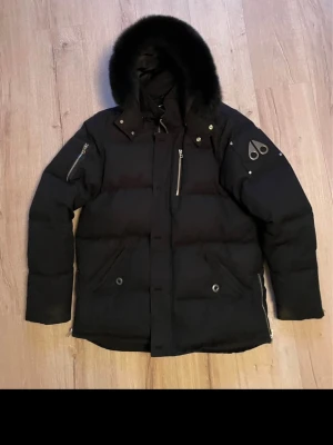 Svart Moose Knuckles pufferjacka - Moose Knuckles med pälskantad. Jackan har dragkedjor på ärmarna, bröstficka med dragkedja och två stora fickor framtill. Kvitto finns vid önskan, storlek Xl men passar även L, inga hål eller fläck tillkommer då jackan är välvårdad.