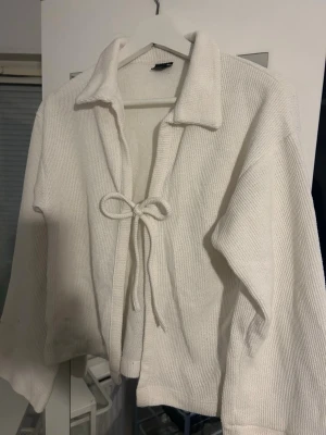 Vit stickad kofta med knyt - Mysig vit stickad kofta med krage och knytband framtill. Tröjan har en relaxed passform och långa ärmar, perfekt för lager på lager. Enkel och stilren design som passar till det mesta.