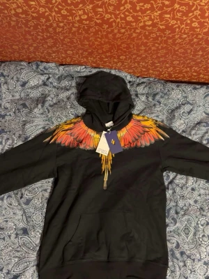 Marcelo Burlon hoodie - COUNTY OF MILAN / Marcelo Burlon Hoodie – Storlek L Säljer en stilren svart hoodie från exklusiva märket County of Milan. Hoodien har en unik och färgstark design runt halsen som liknar fågelfjädrar i nyanser av rött, orange, gult och grått – ett riktigt statement-plagg!  🔹 Storlek: L 🔹 Färg: Svart med flerfärgad grafik 🔹 Skick: Ny med taggar kvar 🔹 Material: Mjuk och bekväm bomullsblandning 🔹 Passform: Normal till något