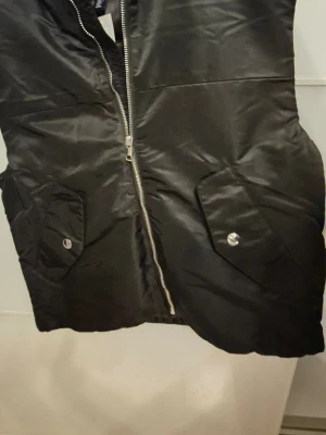 Svart dunväst med dragkedja - Säljer en svart dunväst från H-deluxe med silvrig dragkedja framtill och två stora fickor med lock och knapp. Västen har en clean look och är tillverkad i glansig polyester, perfekt att slänga över en hoodie eller tröja.