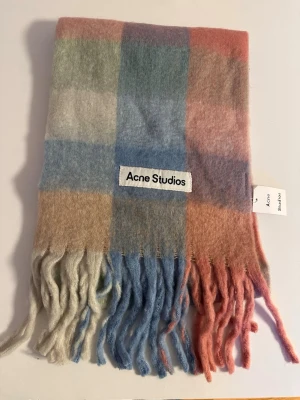 Rutig ullhalsduk från Acne Studios - Supermjuk och fluffig halsduk från Acne Studios i rutigt mönster med pastelliga toner av blått, rosa, beige och grått. Stor logotyp framtill och långa fransar i ändarna. Perfekt accessoar för att lyfta din vinterstil.