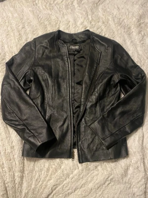 Svart skinnjacka från Classic - Säljer en svart skinnjacka från Classic med stilren design och rundad krage. Jackan har dragkedja framtill och är figursydd för en snygg passform. Perfekt till dig som vill ha en klassisk och tidlös look.
