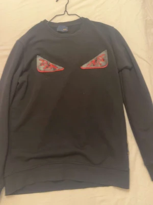 Svart sweatshirt från Fendi med ögon - Är L men sitter som m Cool svart sweatshirt från Fendi med ikoniska ögonapplikationer framtill i svart och rött. Tröjan har rund halsringning och är tillverkad i mjuk bomull. Perfekt för dig som vill sticka ut med en unik detalj på bröstet.