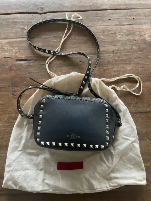 Valentino garvani väska  - Säljer en svarta camera bag från Valentino Garavani med ikoniska silverfärgade nitar runt kanterna och på axelremmen. Har vissa slitna ställen som visas på bilderna men som enkelt skulle kunna gå att fixa. Men väskan i helhet är i ett bra skick. Har dustbag och äktenhetsbevis från vestiaire.