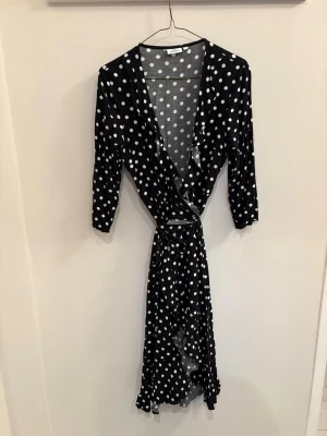 Svart prickig omlottklänning KappAhl - Supersnygg svart omlottklänning från KappAhl med vita prickar och volangkant. Klänningen har trekvartsärm, knyt i midjan och v-ringning. Perfekt för dig som gillar en lekfull och trendig stil.