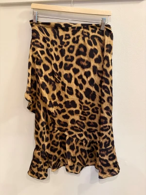 Leopardmönstrad omlottkjol med volang - Säljer en kort omlottkjol med leopardmönster och volangkant. Kjolen har knytband i midjan och är gjord i ett mjukt, glansigt material som ger en snygg och trendig look. Perfekt för dig som vill sticka ut och älskar djurmönster.