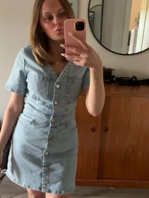 Ljusblå jeansklänning med knappar - Intressekoll på ljusblå jeansklänning med kort ärm och v-ringning. Klänningen har knappar hela vägen framtill, två bröstfickor och två fickor vid höfterna. Perfekt för en avslappnad och trendig look. Skriv för fler bilder eller frågor 🙌