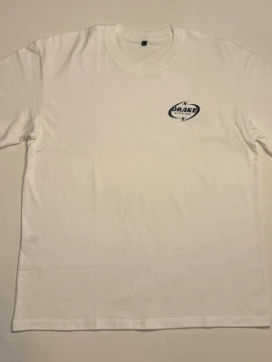 Vit t-shirt från Drake Clothing - Säljer en vit t-shirt från Drake Clothing med tryck på bröstet och ett citat på ryggen. T-shirten har klassisk passform, rund hals och är tillverkad i mjuk bomull. Perfekt för dig som gillar stilrena plagg med coola detaljer. FINNS I L M OCH S