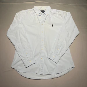 Ljusblå skjorta Polo Ralph Lauren Slim Fit - Klassisk ljusblå skjorta från Polo Ralph Lauren i slim fit-modell. Skjortan har button down-krage, långa ärmar och knappar framtill. Broderad svart logga på bröstet. En perfekt skjorta för en clean och stilren look. Skjortan har en liten missfärgning på insidan av kragen som knappt märks.