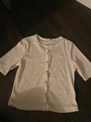 Beige ribbad kofta från H&M - Kortärmad beige kofta från H&M i ribbad bomullsmix. Tröjan har rund halsringning och knappar framtill. 