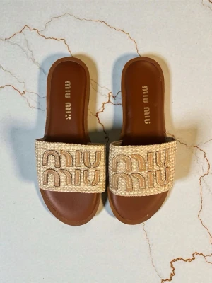 Beige sandaler från Miu Miu - Snygga beige sandaler från Miu Miu med bred flätad rem och broderad logga. Sulan är brun och har en platt form som ger en chill vibe. Perfekta för sommardagar och lätta att matcha med allt. Materialet känns lyxigt och detaljerna är riktigt stilrena.