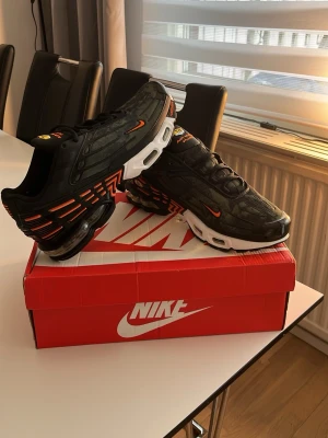 Nike Air Max Plus svart/orange sneakers - Nike Air Max Plus sneakers i svart med orange detaljer och vit sula. Skorna har en tydlig synlig Air-enhet, mesh och syntetmaterial på ovandelen samt coola grafiska linjer i orange längs sidan. Låg modell med snörning och rund tå. Perfekta för dig som gillar streetwear och statement sneakers.