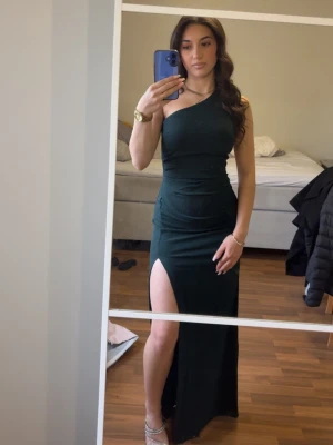 Mörkgrön långklänning med slits - Säljer en elegant mörkgrön långklänning med oneshoulder och hög slits på ena sidan. Klänningen har en tight passform och öppen rygg med dragkedja bak. Perfekt för bal eller festliga tillfällen. Använd en gång. 