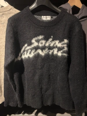 Saint Laurent stickad tröja - Säljer en riktigt fet och eftertraktad svart stickad tröja från Saint Laurent med vit logga i graffiti-stil över bröstet. Tröjan har rund halsringning och långa ärmar. Materialet är fluffigt och mjukt, perfekt för kyliga dagar. Passformen är normal och tröjan är riktigt snygg till jeans. Modellen är 177