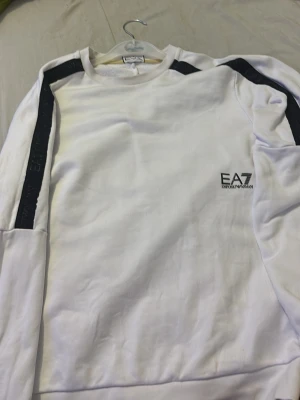 Vit sweatshirt från EA7 Emporio Armani - Säljer en clean vit sweatshirt från EA7 Emporio Armani med svarta detaljer längs axlar och ärmar. Tröjan har rund halsringning, lång ärm och logga på bröstet. Perfekt för dig som gillar stilrena plagg med sportig touch.