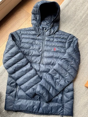 Mörkblå dunjacka från Polo Ralph Lauren - Säljer en mörkblå dunjacka från Polo Ralph Lauren med huva och halv dragkedja. Jackan har quiltad design, lång ärm och den klassiska röda loggan på bröstet. Perfekt för kyliga dagar och riktigt snygg till streetwear-stilen.