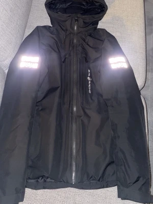 Svart pufferjacka från Sail Racing - Säljer en svart pufferjacka från Sail Racing med huva och reflekterande detaljer på ärmarna. Jackan har dragkedja framtill och är tillverkad i slitstarkt syntetmaterial. Perfekt för kalla dagar och har en clean, sportig look.