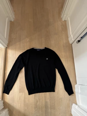 Svart v-ringad tröja Fred Perry L - Svart v-ringad tröja från Fred Perry i storlek L. Liten i storlek sitter som S Klassisk design med vit broderad logga på bröstet och diskret randig detalj i halsringningen. Långärmad och i mjukt material som passar perfekt till lager-på-lager-stil.
