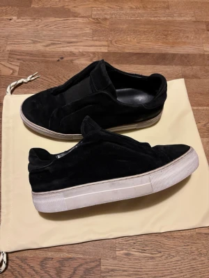 Svarta sneakers från Axel Arigato - Säljer ett par svarta sneakers från Axel Arigato i mocka med vit platt sula. Skorna har minimalistisk design utan snörning och elastiska paneler för enkel påtagning. Märket syns diskret på sidan och tungan. Perfekta för en clean och stilren look. Slitage i hälen av vänstra skon men ingenting man märker medans man har på sig skorna.