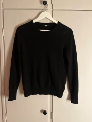 Svart kashmirtröja från Uniqlo - En klassisk svart tröja från Uniqlo i mjuk kashmir. Tröjan har rund halsringning, lång ärm och ribbade muddar vid ärmslut och nederkant. Perfekt för lager på lager under kyliga dagar.